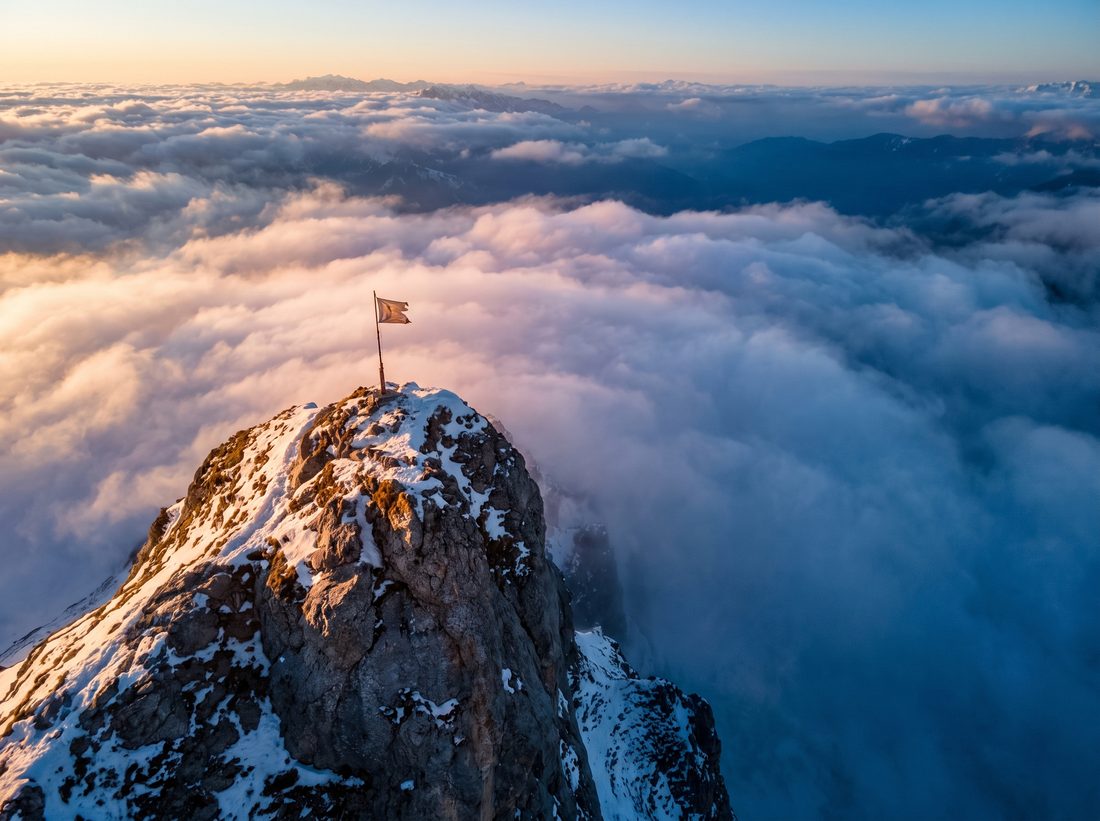Snow-capped mountain peak with a flag at the summit above the clouds